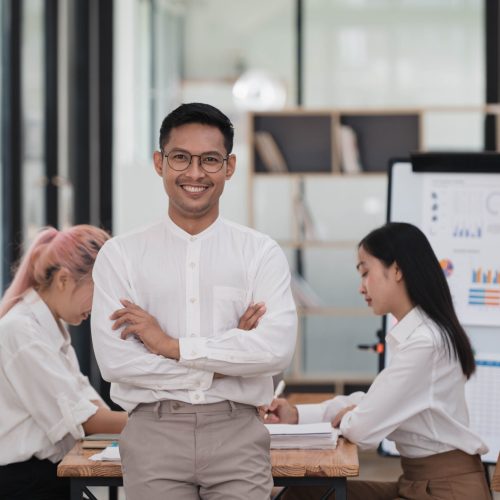 Smiling young Asian businessman standing in the office room with coworkers in the background.